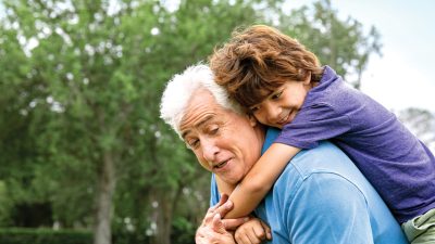 A smiling child rides piggyback on an older man's shoulders outdoors, with green trees behind them.
