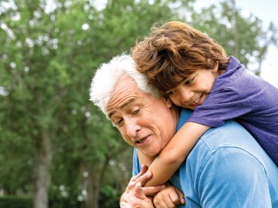 A smiling child rides piggyback on an older man's shoulders outdoors, with green trees behind them.