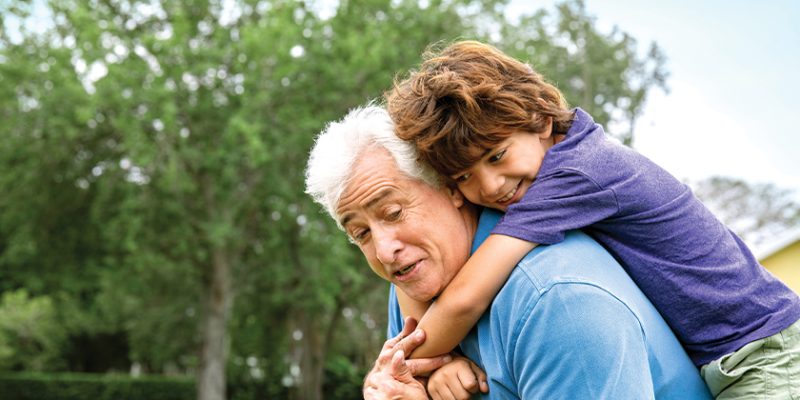 A smiling child rides piggyback on an older man's shoulders outdoors, with green trees behind them.