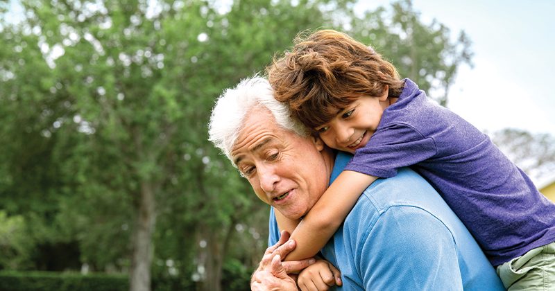 A smiling child rides piggyback on an older man's shoulders outdoors, with green trees behind them.