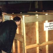 Men in coats gather around a large wooden crate in a dim barn. A sign on the crate reads ORYX from British Zoos to Tunisia.