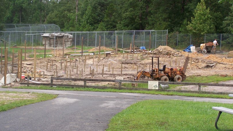 An early zoo habitat under construction: rows of wooden posts set into bare dirt inside a part-built chain-link enclosure, with a small orange backhoe and a worker with a wheelbarrow at the far side