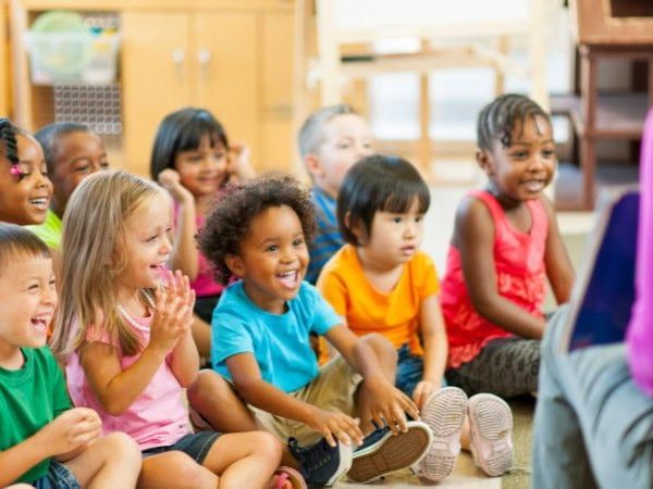 A group of smiling preschool-age children sit cross-legged on the floor in a bright classroom, clapping and looking toward an adult seated in front of them reading aloud