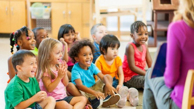 A group of smiling preschool-age children sit cross-legged on the floor in a bright classroom, clapping and looking toward an adult seated in front of them reading aloud