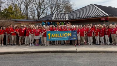 A large group of volunteers in red shirts stands outside a zoo building holding a banner reading Celebrating 1 Million Volunteer Hours.