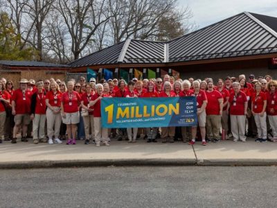 A large group of volunteers in red shirts stands outside a zoo building holding a banner reading Celebrating 1 Million Volunteer Hours.
