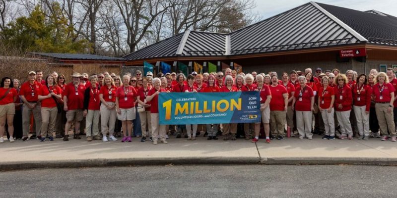 A large group of volunteers in red shirts stands outside a zoo building holding a banner reading Celebrating 1 Million Volunteer Hours.