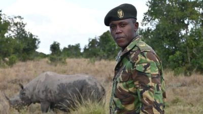 An Ol Pejeta Conservancy ranger in camouflage uniform and beret standing in open grassland, looking toward the camera, with a rhino grazing in the grass behind him and acacia scrub on the horizon.
