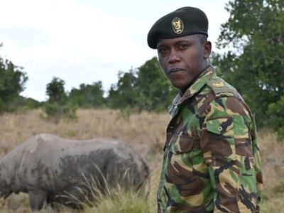 An Ol Pejeta Conservancy ranger in camouflage uniform and beret standing in open grassland, looking toward the camera, with a rhino grazing in the grass behind him and acacia scrub on the horizon.
