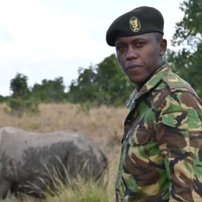 An Ol Pejeta Conservancy ranger in camouflage uniform and beret standing in open grassland, looking toward the camera, with a rhino grazing in the grass behind him and acacia scrub on the horizon.