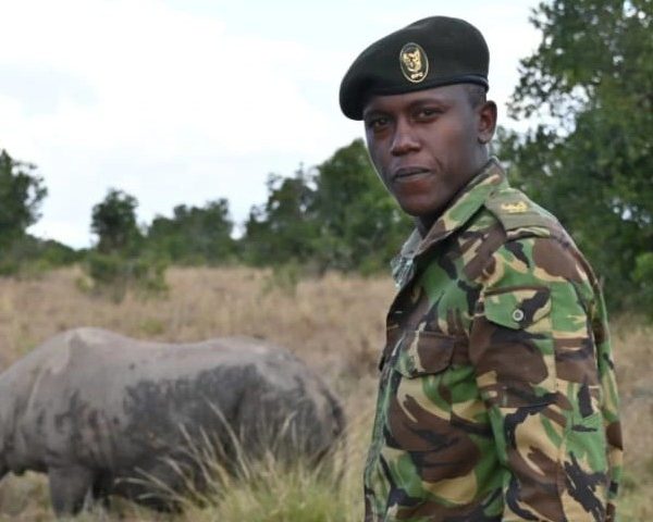 An Ol Pejeta Conservancy ranger in camouflage uniform and beret standing in open grassland, looking toward the camera, with a rhino grazing in the grass behind him and acacia scrub on the horizon.