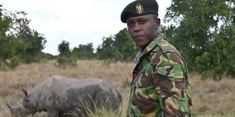 An Ol Pejeta Conservancy ranger in camouflage uniform and beret standing in open grassland, looking toward the camera, with a rhino grazing in the grass behind him and acacia scrub on the horizon.