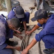Three young trainees in blue mechanic's overalls and caps crouching on the ground beside a pickup truck, working together on a leaf-spring assembly with hand tools.