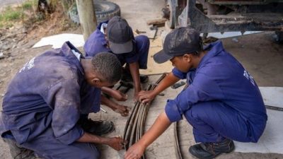 Three young trainees in blue mechanic's overalls and caps crouching on the ground beside a pickup truck, working together on a leaf-spring assembly with hand tools.