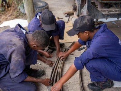 Three young trainees in blue mechanic's overalls and caps crouching on the ground beside a pickup truck, working together on a leaf-spring assembly with hand tools.