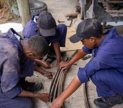 Three young trainees in blue mechanic's overalls and caps crouching on the ground beside a pickup truck, working together on a leaf-spring assembly with hand tools.