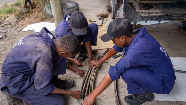 Three young trainees in blue mechanic's overalls and caps crouching on the ground beside a pickup truck, working together on a leaf-spring assembly with hand tools.