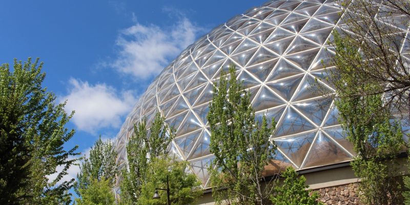 The glass geodesic roof of the Desert Dome at Omaha's Henry Doorly Zoo and Aquarium rises above green trees under a blue sky.