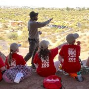 Two City of Phoenix park rangers in uniform stand outdoors with a group of children, pointing toward desert vegetation under a bright sky.