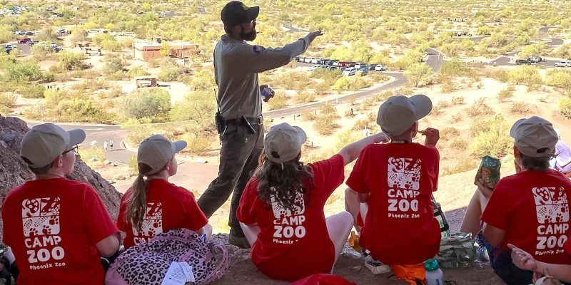Two City of Phoenix park rangers in uniform stand outdoors with a group of children, pointing toward desert vegetation under a bright sky.