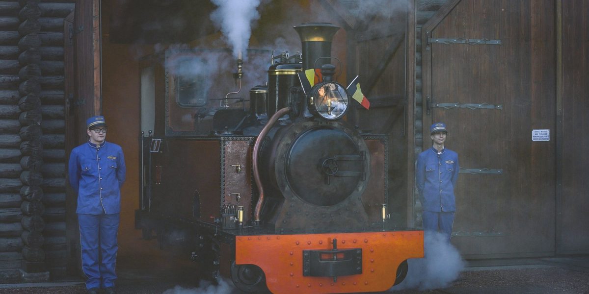 A small restored steam locomotive with an orange buffer beam stands under steam in the open doorway of a timber engine shed at Pairi Daiza, a Belgian flag mounted beside its headlamp, with a member of staff in a blue railway uniform and cap standing on either side of it.