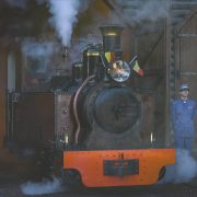 A small restored steam locomotive with an orange buffer beam stands under steam in the open doorway of a timber engine shed at Pairi Daiza, a Belgian flag mounted beside its headlamp, with a member of staff in a blue railway uniform and cap standing on either side of it.