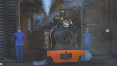 A small restored steam locomotive with an orange buffer beam stands under steam in the open doorway of a timber engine shed at Pairi Daiza, a Belgian flag mounted beside its headlamp, with a member of staff in a blue railway uniform and cap standing on either side of it.