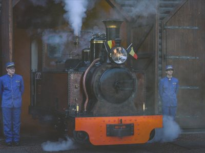 A small restored steam locomotive with an orange buffer beam stands under steam in the open doorway of a timber engine shed at Pairi Daiza, a Belgian flag mounted beside its headlamp, with a member of staff in a blue railway uniform and cap standing on either side of it.