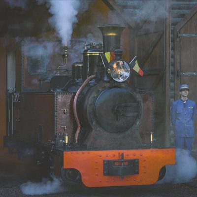 A small restored steam locomotive with an orange buffer beam stands under steam in the open doorway of a timber engine shed at Pairi Daiza, a Belgian flag mounted beside its headlamp, with a member of staff in a blue railway uniform and cap standing on either side of it.