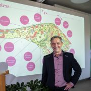 A man in a dark jacket stands in front of a projected site map of the Wilhelma showing labeled future projects.