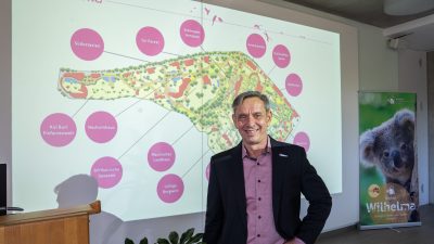 A man in a dark jacket stands in front of a projected site map of the Wilhelma showing labeled future projects.