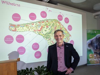 A man in a dark jacket stands in front of a projected site map of the Wilhelma showing labeled future projects.