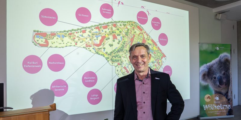 A man in a dark jacket stands in front of a projected site map of the Wilhelma showing labeled future projects.