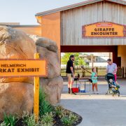 A sign reading Helmerich Meerkat Exhibit stands beside a boulder wall on a new paved path at Tulsa Zoo, with the timber-framed Hardesty Family Foundation Giraffe Encounter building and several visitors behind it.
