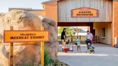 A sign reading Helmerich Meerkat Exhibit stands beside a boulder wall on a new paved path at Tulsa Zoo, with the timber-framed Hardesty Family Foundation Giraffe Encounter building and several visitors behind it.