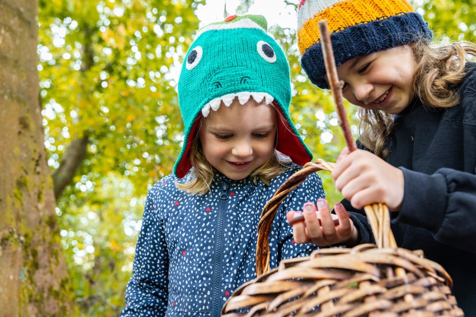 Two children in woodland in autumn, one in a green dinosaur hat holding a wicker basket and the other in a yellow and navy bobble hat holding a long stick, both looking down into the basket.