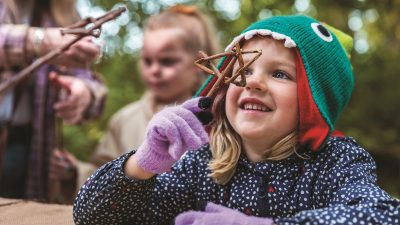 A young child in a green knitted hat shaped like a fish holds a cluster of twigs up to her face, smiling, with an adult and another child blurred behind her in autumn woodland.