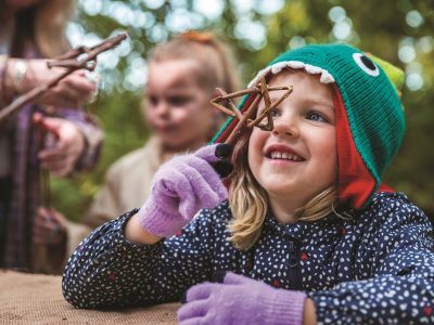 A young child in a green knitted hat shaped like a fish holds a cluster of twigs up to her face, smiling, with an adult and another child blurred behind her in autumn woodland.