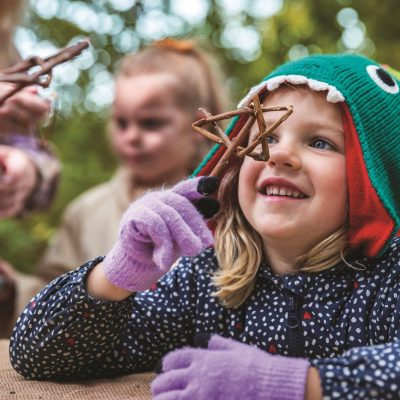 A young child in a green knitted hat shaped like a fish holds a cluster of twigs up to her face, smiling, with an adult and another child blurred behind her in autumn woodland.