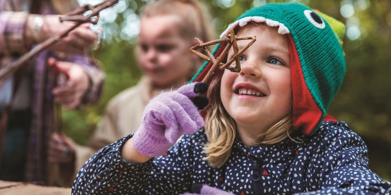 A young child in a green knitted hat shaped like a fish holds a cluster of twigs up to her face, smiling, with an adult and another child blurred behind her in autumn woodland.