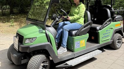 A zoo staff member sits at the wheel of a small green electric vehicle with an open side and a boarding ramp.
