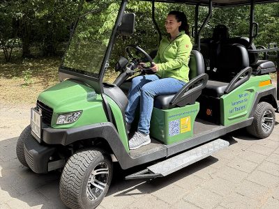 A zoo staff member sits at the wheel of a small green electric vehicle with an open side and a boarding ramp.