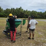 Four people on a grass airstrip fringed by coconut palms assemble a large green conical hopper mounted on a wheeled frame, part of the equipment for the Alphonse Atoll invasive species eradication.