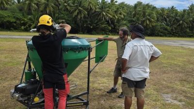 Four people on a grass airstrip fringed by coconut palms assemble a large green conical hopper mounted on a wheeled frame, part of the equipment for the Alphonse Atoll invasive species eradication.