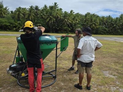 Four people on a grass airstrip fringed by coconut palms assemble a large green conical hopper mounted on a wheeled frame, part of the equipment for the Alphonse Atoll invasive species eradication.