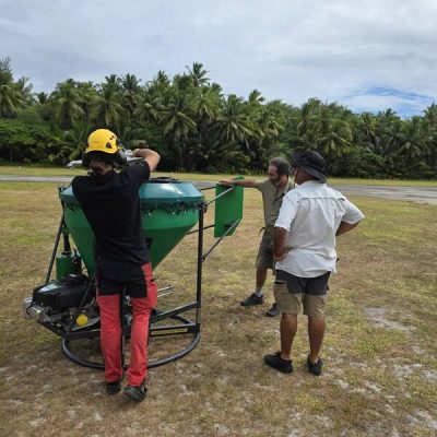 Four people on a grass airstrip fringed by coconut palms assemble a large green conical hopper mounted on a wheeled frame, part of the equipment for the Alphonse Atoll invasive species eradication.