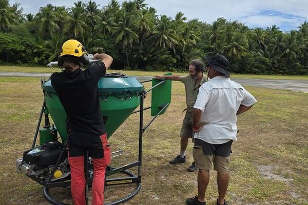Four people on a grass airstrip fringed by coconut palms assemble a large green conical hopper mounted on a wheeled frame, part of the equipment for the Alphonse Atoll invasive species eradication.