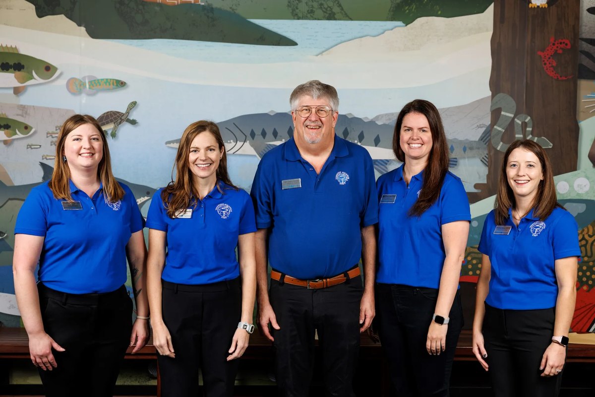 Five Tennessee Aquarium animal health staff in blue polo shirts stand in a row in front of a painted mural of river fish, a turtle and a salamander.
