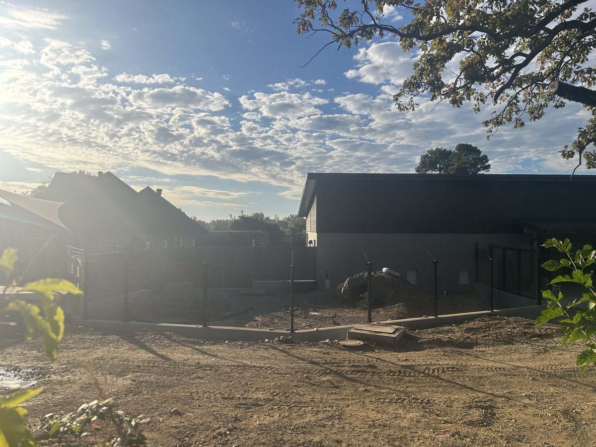 Black exhibit fencing and upright posts enclose a bare earth yard beside a dark-roofed new building at the Little Rock Zoo, with construction tracks visible in the dirt and morning sun behind the trees.