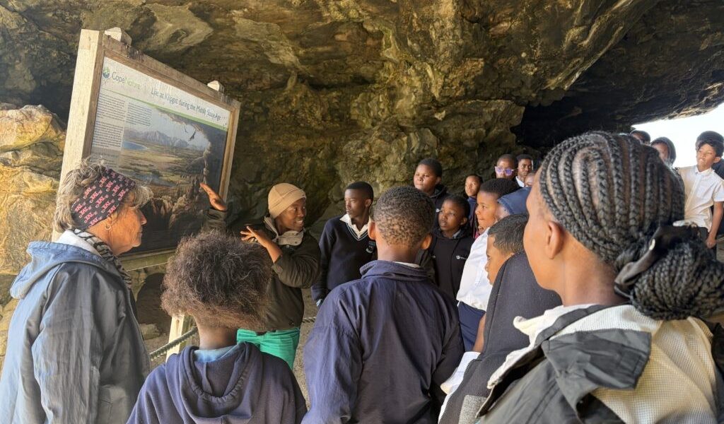 A group of school learners stands inside Klipgat Cave around a CapeNature interpretive board headed Life at Klipgat during the Middle Stone Age.